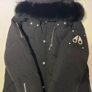 Svart vinterjacka från Moose Knuckles - Säljer en svart dunjacka från Moose Knuckles med fluffig fuskpäls runt huvan, silvriga dragkedjor och knappar samt logga på ärmen. Jackan har flera fickor och en riktigt schysst oversized look. Perfekt för kalla vinterdagar.