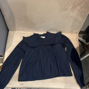 Mörkblå blus med volang från Gina Tricot - Supersöt mörkblå blus från Gina Tricot i storlek 146/152. Blusen har långa ärmar, volangdetaljer vid halsringningen och fina broderade partier över bröstet och på ärmarna. Perfekt för dig som gillar en romantisk och trendig stil.