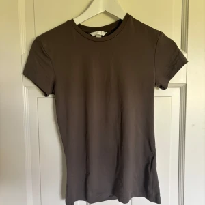 Brun basic t-shirt från H&M - En stilren brun t-shirt från H&M i storlek S. Modellen är klassisk med rund halsringning och korta ärmar. Tillverkad i mjuk bomull som känns skön mot huden. Perfekt att matcha med jeans eller kjol för en enkel och clean look. Aldrig använd, men är utan lapp. 