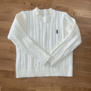 Vit stickad tröja från Ralph Lauren - Säljer en vit kabelstickad tröja från Ralph Lauren med klassisk rund hals och ribbade muddar. Tröjan har det ikoniska Polo-logot broderat i svart på bröstet. Perfekt för dig som gillar stilrena och tidlösa plagg.