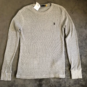Grå långärmad tröja Polo Ralph Lauren - Grå långärmad tröja från Polo Ralph Lauren med klassisk svart broderad logga på bröstet. Tröjan är ribbstickad och har rund halsringning. Perfekt för lager-på-lager och chill vardagsstil.