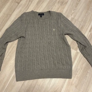 Grå kabelstickad tröja Polo Ralph Lauren - Kabelstickad grå tröja från Polo Ralph Lauren med klassisk rund hals och broderad logga på bröstet. Tröjan har lång ärm och ribbade muddar för en snygg passform. Perfekt för lager-på-lager under kyliga dagar. Är 180 lång å den är lite för liten så skulle nog passa 170-175 