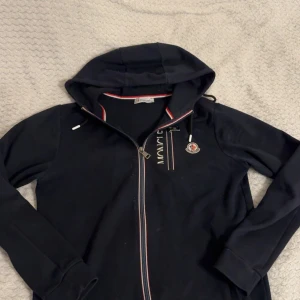 Marine blå hoodie från Moncler med dragkedja - Svart hoodie från Moncler med hel dragkedja och huva. Tricolore-detaljer vid dragkedjan och insidan av kragen. Klassisk Moncler-logga på bröstet och snörning i huvan. Perfekt för en stilren och sportig look.