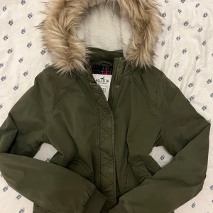 Vintage olivgrön jacka med fuskpäls från Hollister - Snygg vintage khaki / olivgrön bomberjacka från Hollister med huva och fuskpäls. Insidan har röd- och grönrutigt foder och mjuk teddy vid huvan. Jackan har dragkedja och muddar vid ärmslut och midja. Perfekt för kyliga dagar och riktigt skön att bära. Jag brukar har s och den är lite oversized på mig hade varit perfekt för att ha en hoodie under typ. 