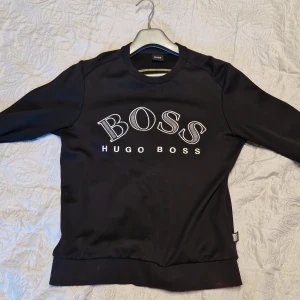 Svart sweatshirt från Hugo Boss - Svart sweatshirt från Hugo Boss med stor vit BOSS-logga framtill. Tröjan har rund halsringning, långa ärmar och ribbade muddar. Tillverkad i mjuk bomullsmix som är skön att bära. Perfekt för en stilren och avslappnad look.