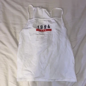 Vit linne-topp med tryck från H&M - Vit linne-topp från H&M med tunna axelband och tryck på bröstet med texten '1994 USA' i svart och rött. Toppen är i mjuk bomull och har en enkel, sportig look som passar perfekt till sommaren.