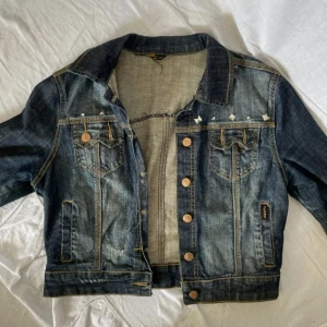 Jeansjacka med nitar och slitningar - Säljer en blå jeansjacka med coola slitningar och silvriga nitar längs axlarna. Klassisk modell med bröstfickor, knappar framtill och kontrastsömmar. Perfekt för dig som vill ha en edgy och trendig look.