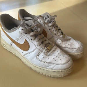 Nike Air Force 1 vita sneakers - Nike Air Force 1 sneakers i vitt skinn med bruna detaljer och swoosh. Klassisk siluett med perforerad tå, snörning och platt sula. Insidan har meshfoder och gröna innersulor. Perfekta för dig som gillar retrovibbar och streetstyle.