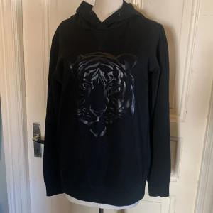 Svart hoodie med tigertryck - Svart hoodie med snyggt tigertryck i blankt svart framtill. Tröjan har huva med snörning och långa ärmar. Perfekt för dig som gillar streetstyle och vill sticka ut med ett coolt motiv.