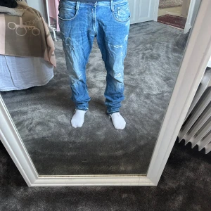 Replay Anbass blå jeans med slitningar - Säljer ett par Replay Anbass jeans i blå tvätt med coola slitningar och lätt blekta detaljer. Modellen har normal passform och raka ben, klassisk femficksdesign och läderpatch bak i midjan. Perfekt för dig som gillar en avslappnad streetstil.