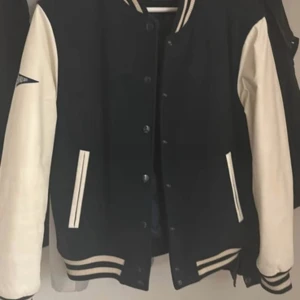 Svart och beige bomberjacka Superdry - Snygg svart och beige bomberjacka från Superdry med klassiska kontrastärmar i beige skinn och svart kropp i ullmix. Jackan har ribbade muddar med ränder, knappar framtill och två fickor. Perfekt för dig som gillar sportig och retro stil. Helt ny, oanvänd.