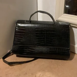 Svart axelväska i krokopräglat skinn - Stilren svart axelväska med krokodilpräglat mönster och både handtag och axelrem. Väskan har en klassisk, fyrkantig form och är tillverkad i glansigt skinnliknande material. Perfekt för dig som vill ha en elegant och trendig accessoar.