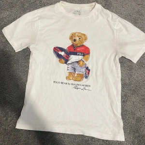Vit Polo Bear t-shirt från Ralph Lauren - Vit t-shirt från Polo Ralph Lauren med ikoniskt Polo Bear-tryck på framsidan. Klassisk rund hals och korta ärmar. Tillverkad i mjuk bomull som känns skön mot huden. Perfekt för dig som gillar stilrena plagg med en lekfull twist.