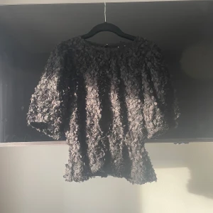 Svart blommig blus från Gina Tricot - Supersnygg svart blus från Gina Tricot med 3D-blommor över hela tyget. Blusen har korta puffärmar och rund halsringning. Materialet är spetsigt och ger en cool struktur. Perfekt för dig som vill sticka ut med en unik topp!