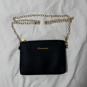 Svart quiltad axelväska - Snygg svart axelväska med quiltat sicksackmönster och guldfärgad kedja. Väskan är i skinnimitation och har dragkedja upptill. Perfekt storlek för det viktigaste och ger en trendig touch till din outfit.
