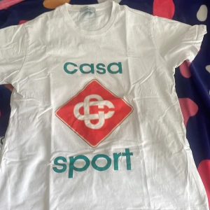 Vit Casa Sport t-shirt från Casablanca - Vit t-shirt från Casablanca med tryck i turkos och rött framtill där det står 'casa sport' och en grafisk logga. Klassisk passform med rund hals och korta ärmar. Tillverkad i mjuk bomull, perfekt för en sportig och clean look. Inte använd mycket, den är ny!