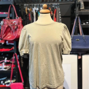 Beige/grön topp med puffärm från Vero Moda - Snygg beige/grön topp från Aware by Vero Moda i 100% bomull. Modellen har puffiga korta ärmar och rund halsringning, vilket ger en trendig och avslappnad look. Perfekt att styla med jeans eller kjol för en enkel men cool outfit.
