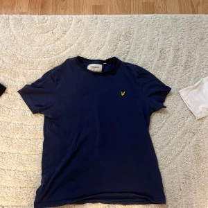 Marinblå t-shirt från Lyle & Scott - Marinblå t-shirt från Lyle & Scott med klassisk rund hals och gul broderad logga på bröstet. Tillverkad i ekologisk bomull för en skön och avslappnad känsla. Perfekt basic-plagg med snygg passform.