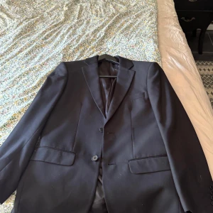 Svart kostym Lindbergh White slim fit - Säljer en svart kostym från Lindbergh White i slim fit-modell. Kavajen har två knappar, klassiska fickor med lock och slät design. Kostymen är tillverkad av återvunna fibrer och har en stilren, modern look. Perfekt för dig som gillar minimalistisk stil och snygg passform.