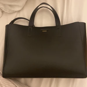 Svart handväska från Wera i skinn - Stilren svart handväska från Wera i slätt skinn. Väskan har två handtag, avtagbar axelrem och magnetlås. Insidan är rymlig med fack för småsaker. Perfekt för dig som gillar minimalistisk och klassisk design.