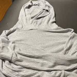 Ljusgrå ribbad hoodie med huva - Mysig ljusgrå hoodie med ribbad struktur och huva. Tröjan har långa ärmar och är tillverkad i ett mjukt material som passar perfekt för kyligare dagar. Enkel och stilren design som är lätt att matcha med andra plagg.