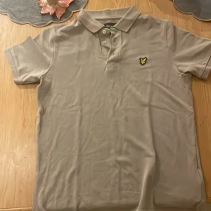 Beige pikétröja från Lyle & Scott - Snygg beige pikétröja från Lyle & Scott med klassisk krage, korta ärmar och knäppning framtill. Tröjan har den ikoniska gula örnloggan broderad på bröstet. Perfekt för en clean och stilren look.