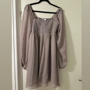 Lila kortklänning med puffärmar - Supersöt lila kortklänning med transparenta puffiga långärmar och smockad överdel. Klänningen har små prickar i tyget och en flowy kjol som ger en romantisk vibe. Perfekt för dig som gillar en feminin och trendig stil.