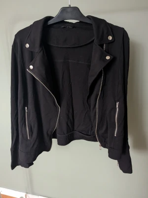 Svart bikerinspirerad jacka - Säljer en svart jacka med bikerstil, tillverkad i mjukt bomullsmaterial. Jackan har silvriga dragkedjor framtill och på fickorna, samt tryckknappar på kragen. Långärmad och med coola detaljer som ger en edgy look.