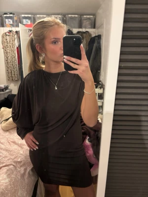 Brun  kortklänning - Säljer en svart kortklänning med cape design och en ärm. Klänningen har en lösare passform upptill och smalnar av nedtill. Materialet  mjukt och stretchigt.