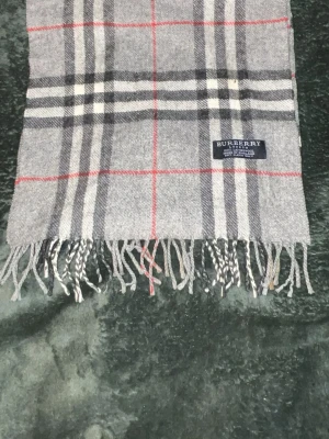 Grå rutig halsduk från Burberry - Klassisk grå halsduk från Burberry i 100% lammull. Den har det ikoniska rutiga mönstret i grått, svart, vitt och rött samt fransar i ändarna. Perfekt accessoar för att lyfta din outfit med en tidlös brittisk vibe.