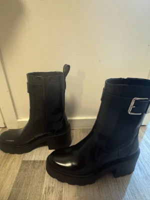 Svarta boots med grov sula och spänne - Säljer ett par svarta boots i glansigt skinn med chunky klack och grov sula. Skorna har ett snyggt spänne i silver på sidan och dragkedja på insidan för enkel påtagning. Perfekta för dig som vill ha en edgy look till hösten eller vintern.