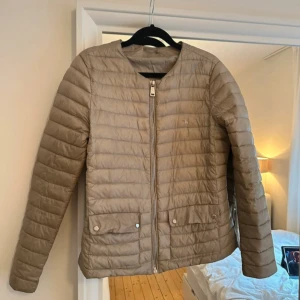 Beige pufferjacka från Polo Ralph Lauren - Säljer en beige pufferjacka från Polo Ralph Lauren med diskret logga på bröstet. Jackan har raka, quiltade paneler, dragkedja framtill och två fickor med knappstängning. Långärmad modell i lätt och smidigt syntetmaterial, perfekt för kyliga dagar.