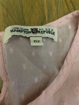 Ljusrosa prickig blus Hampton Republic - Söt ljusrosa blus från Hampton Republic i storlek 152. Blusen är ärmlös och har ett diskret prickigt mönster samt dekorativa sömmar framtill. Tillverkad i ett tunt och luftigt bomullstyg, perfekt för varma dagar.