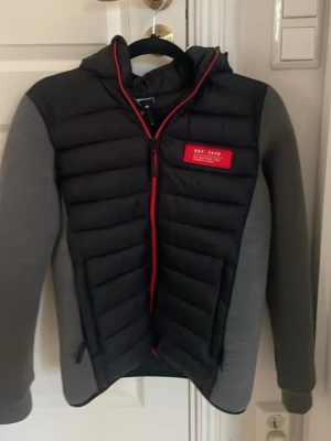 Svart pufferjacka från Brunotti - Säljer en svart och grå cardigan från Brunotti med huva och röda detaljer. Jackan har quiltad framsida, röd dragkedja och en röd patch på bröstet. Ärmarna är grå och materialet är syntet. Perfekt för vinter vår å höst dagar.