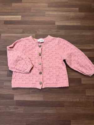 Rosa stickad kofta - Endast använd fåtal gånger, helt utan anmärkningar. Supersöt rosa stickad kofta från Newbie storlek 68 (4-6mån). Har fina hålmönster och träknappar framtill samt puffärmar och ribbade muddar vid ärmslut och nederkant. Perfekt för lager på lager under kyliga dagar.