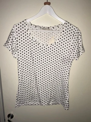 Vit prickig t-shirt från 157 - Snygg vit t-shirt från 157 med svarta prickar över hela plagget. Klassisk rund halsringning och korta ärmar. Tillverkad i mjuk bomull som känns skön mot huden. Perfekt pyjamas tröja eller hemma klädsel. Helt ny med prislappen kvar
