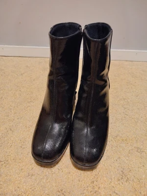 Svarta stövletter med klack och dragkedja - Säljer ett par svarta stövletter i glansigt skinnimitation med rund tå och chunky klack. Skorna har dragkedja på insidan för enkel påtagning och en stilren look som passar till många outfits. Använd 1-2 gånger. Köpta på H&M, storlek 38. Hämtas/lämnas runt Stockholm city/solna helst.