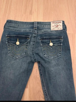 True Religion Joey bootcut jeans blå - Säljer ett par klassiska True Religion Joey jeans i blå tvätt med bootcut passform. Byxorna har låg midja, dekorativa ficklock bak med guldfärgade knappar och kontrastsömmar. Tillverkade i slitstark denim med snygga detaljer och ikonisk etikett bak.