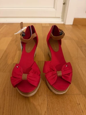 Röda sandaler med kilklack från Tommy Hilfiger - Snygga röda sandaler från Tommy Hilfiger med kilklack i rep och bruna ankelremmar. Skorna har öppen tå, stor rosett framtill och små Hilfiger-loggor på rosetterna. Perfekta för sommaren och ger en färgklick till din outfit.