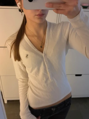 Henley Abercrombie & Fitch långärmad - Vit långärmad henley topp från Abercrombie & Fitch med v-ringning och knappar framtill. Storlek S! Toppens material är mjuk bomull och den har en liten broderad älg-logga på bröstet. Perfekt till jeans eller kjol för en clean och enkel look.