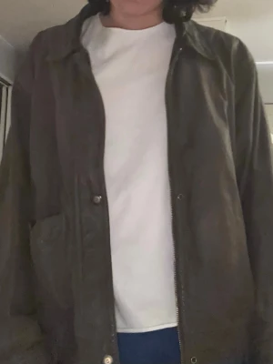 Mörkbrun läderjacka - Säljer en mörkbrun läderjackan med klassisk krage och dragkedja framtill. Jackan har två fickor på sidorna och ribbade muddar vid ärmslut och nederkant. Insidan har ett diskret mönster. Perfekt för dig som vill ha en stilren och tidlös look.