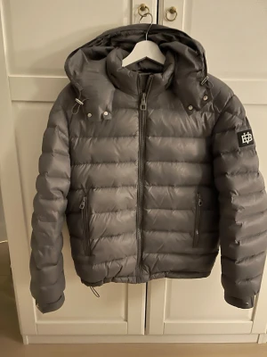 Grå dunjacka från Bvaldi med huva stl M - Säljer en grå pufferjacka från Bvaldi med avtagbar huva och dragkedja framtill. Jackan har två fickor med dragkedja och en snygg logga på ärmen. Fylld med 90% vitt ankdun och 10% fjäder för extra värme. Perfekt för kalla dagar. Storlek M.