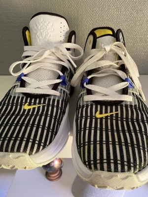 Nike Lebrone 6 - Säljer ett par feta Nike Lebron 6 med svartvitt rutmönster, gula detaljer och blå snörningshål. Skorna har vit mesh, svart sula med blå air-bubbla och coolt broderat lejon på plösen. Perfekta för dig som vill sticka ut med din stil.