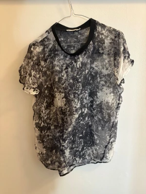 Svartvit transparent blus - Säljer en luftig och transparent blus med abstrakt svartvitt mönster. Blusen har rund halsringning med svart kant och korta ärmar. Materialet är tunt och skönt, perfekt för lager på lager eller till ett par jeans.