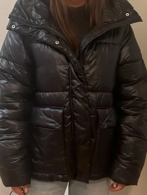 Svart pufferjacka indiska - Säljer en svart pufferjacka med glansig finish och stora framfickor. Snygg krage som är vikt ner på bildern. Storlek xs men sitter som S-M. Märke: indiska 
