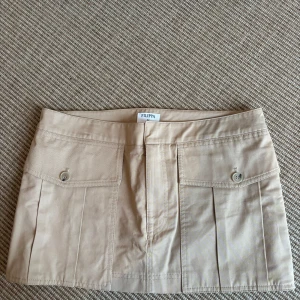 Beige kortkjol från Filippa - Säljer en beige kortkjol från Filippa med två fickor framtill som har knappdetaljer. Kjolen har en rak passform och är gjord i ett mjukt bomullsmaterial. Perfekt för dig som gillar minimalistisk och clean stil.