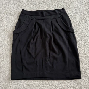 Svart pennkjol från KappAhl - Säljer en svart pennkjol från KappAhl i storlek 36/38. Kjolen har hög midja, dekorativa fickor bak och veck framtill som ger en snygg siluett. Dragkedja bak för enkel påklädning. Perfekt till en stilren look.