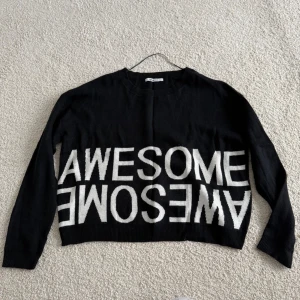 Svart stickad tröja med text från Gina Tricot - Cool svart stickad tröja från Gina Tricot i storlek L. Tröjan har vit text 'AWESOME' både rättvänt och upp och ner över magen. Långärmad modell med ribbade muddar och rund hals. Perfekt för dig som vill ha en statement piece i garderoben.