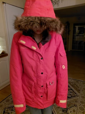 Rosa vinterjacka från Scott med fuskpäls - Riktigt snygg rosa vinterjacka från Scott med huva och fuskpäls. Jackan har reflexdetaljer på ryggen, flera fickor och quiltat foder. Perfekt för kalla dagar och har en sportig vibe. Stängs med dragkedja och knappar. Märke synligt på bröstet och ärmen.
