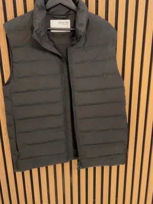 Svart dunväst från Selected Homme - Säljer en svart dunväst från Selected Homme med quiltad design och hög krage. Västen har dragkedja framtill och en stilren, enkel look som passar till många outfits. Perfekt för lager-på-lager under kyligare dagar.